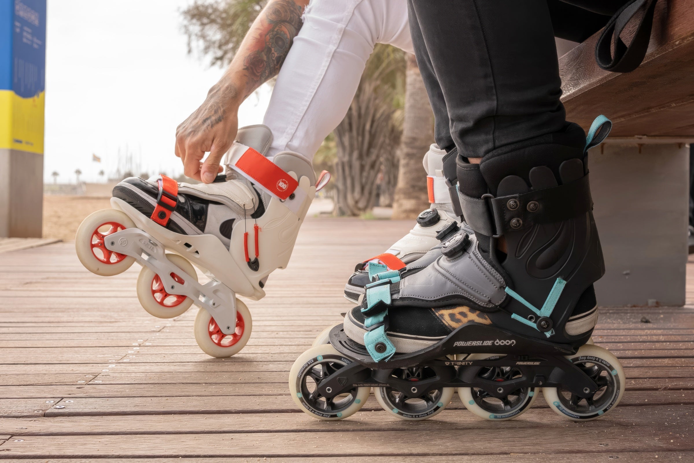 Close-up of two skaters on a wooden boardwalk—one adjusting a white and red tri-wheel skate, while another wears black and gray Powerslide skates with teal straps and a black ankle support.